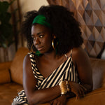 Woman in a striped dress, green headband with gold jewelry sitting on a brown couch.