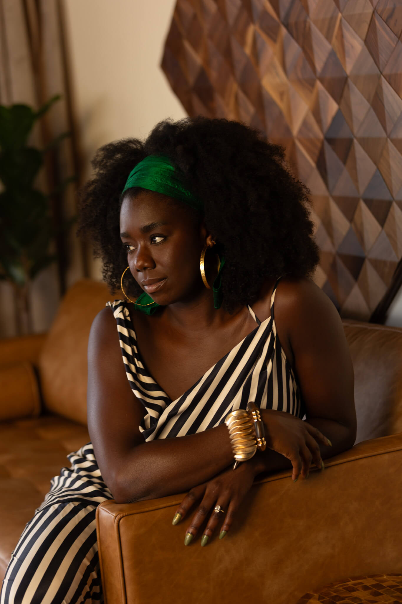 Woman in a striped dress, green headband with gold jewelry sitting on a brown couch.