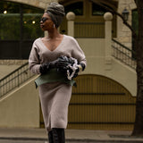 Woman in a beige sweater and black gloves holding a green clutch against a building background