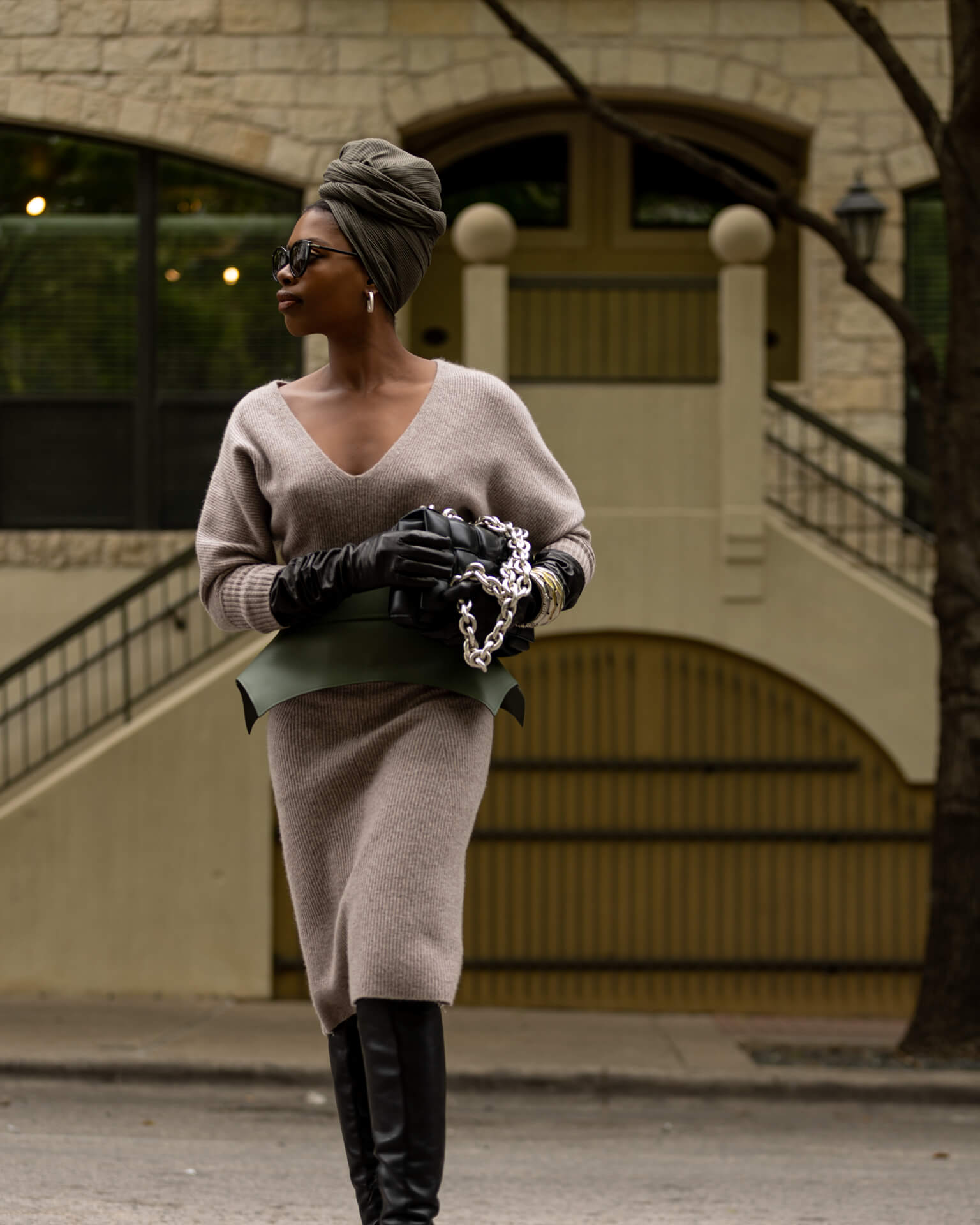 Woman in a beige sweater and black gloves holding a green clutch against a building background