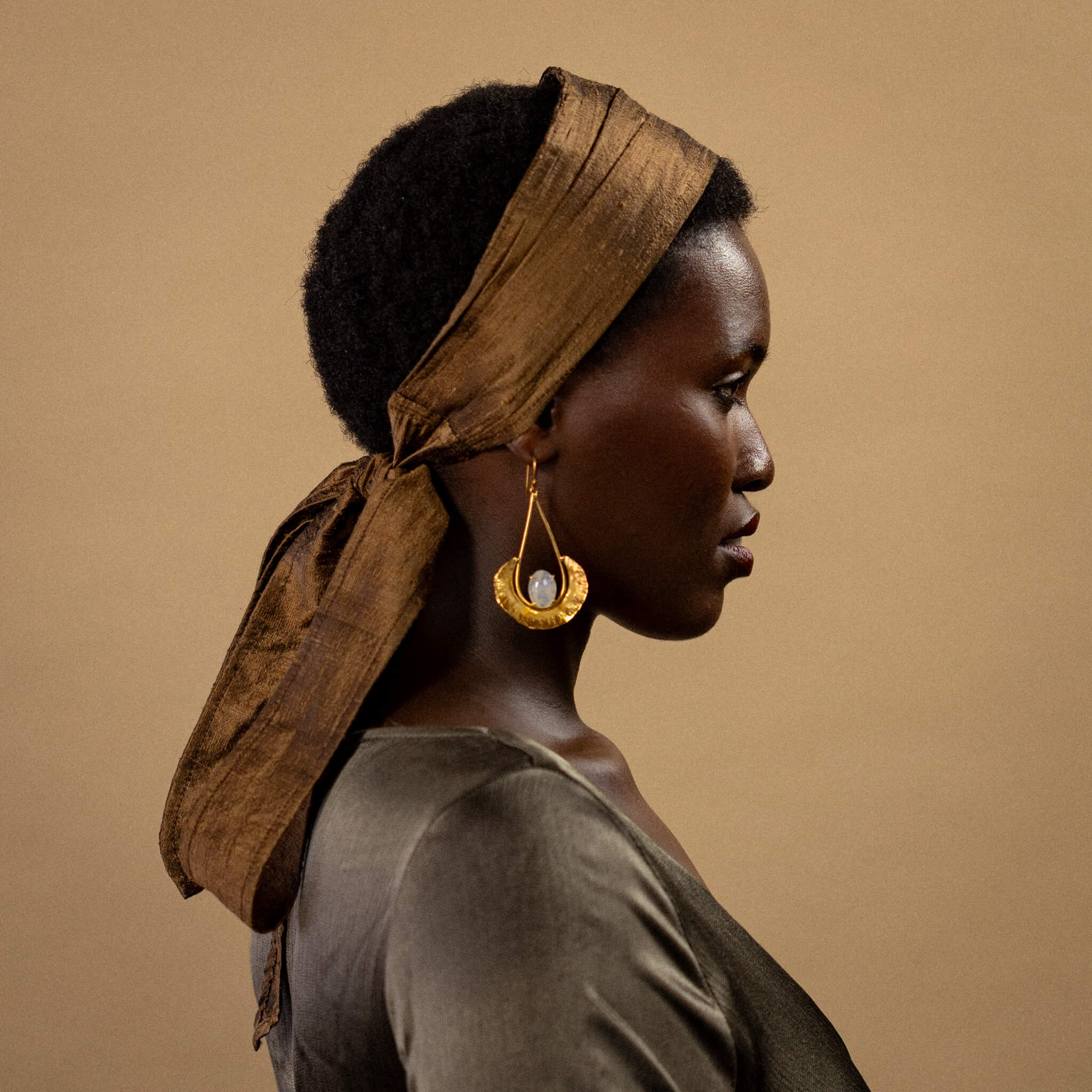 Woman wearing a brown headscarf and gold earrings against a beige background
