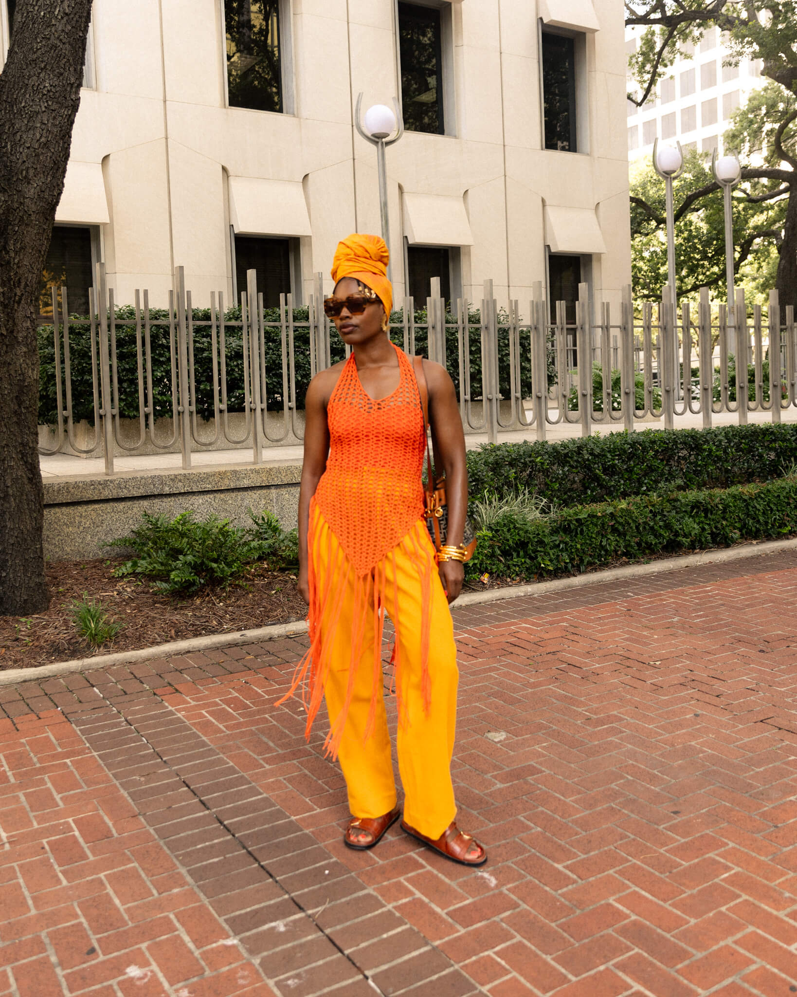 Woman in bright orange outfit standing on a brick sidewalk with a building in the background