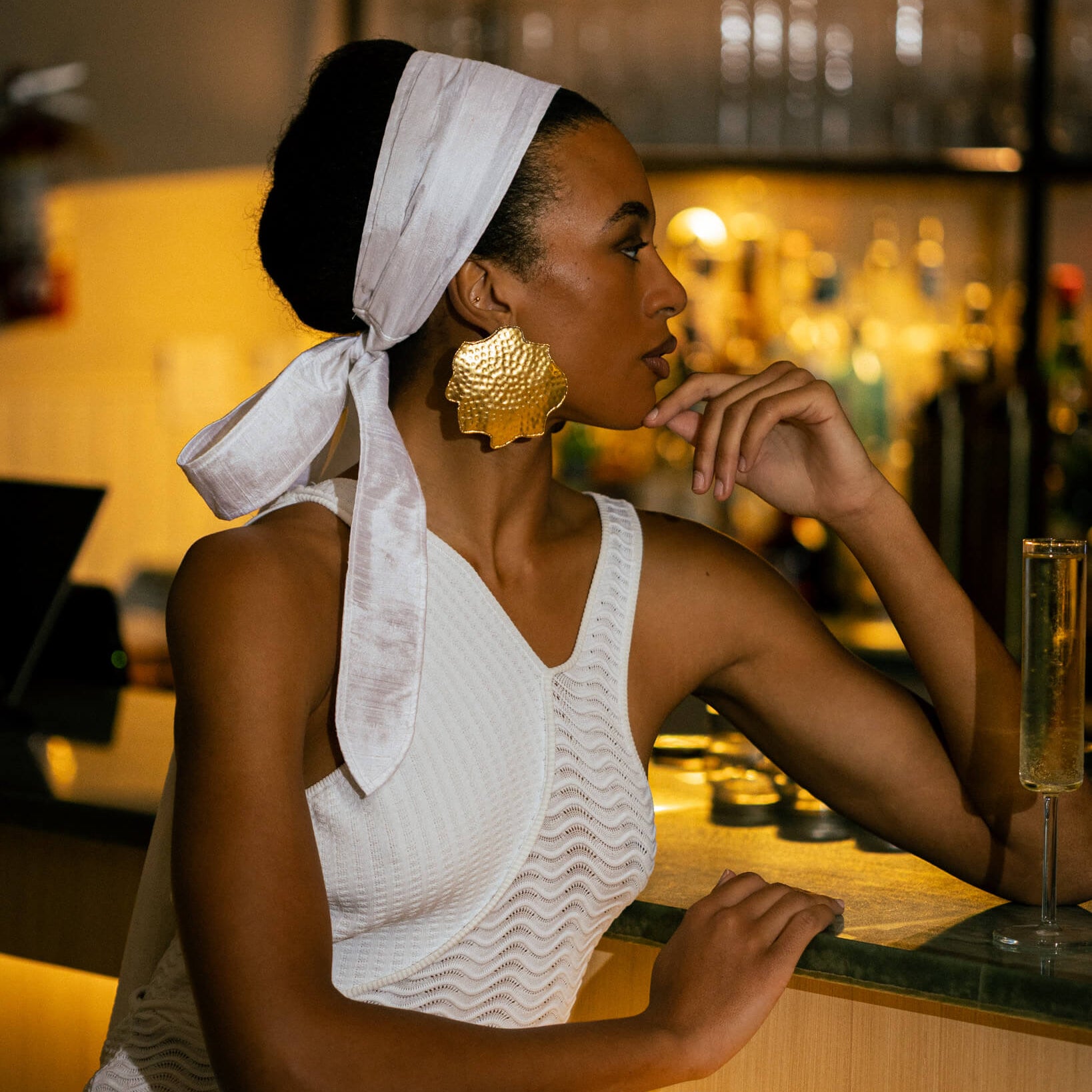 Woman sitting at a bar with a white headscarf and gold earrings.