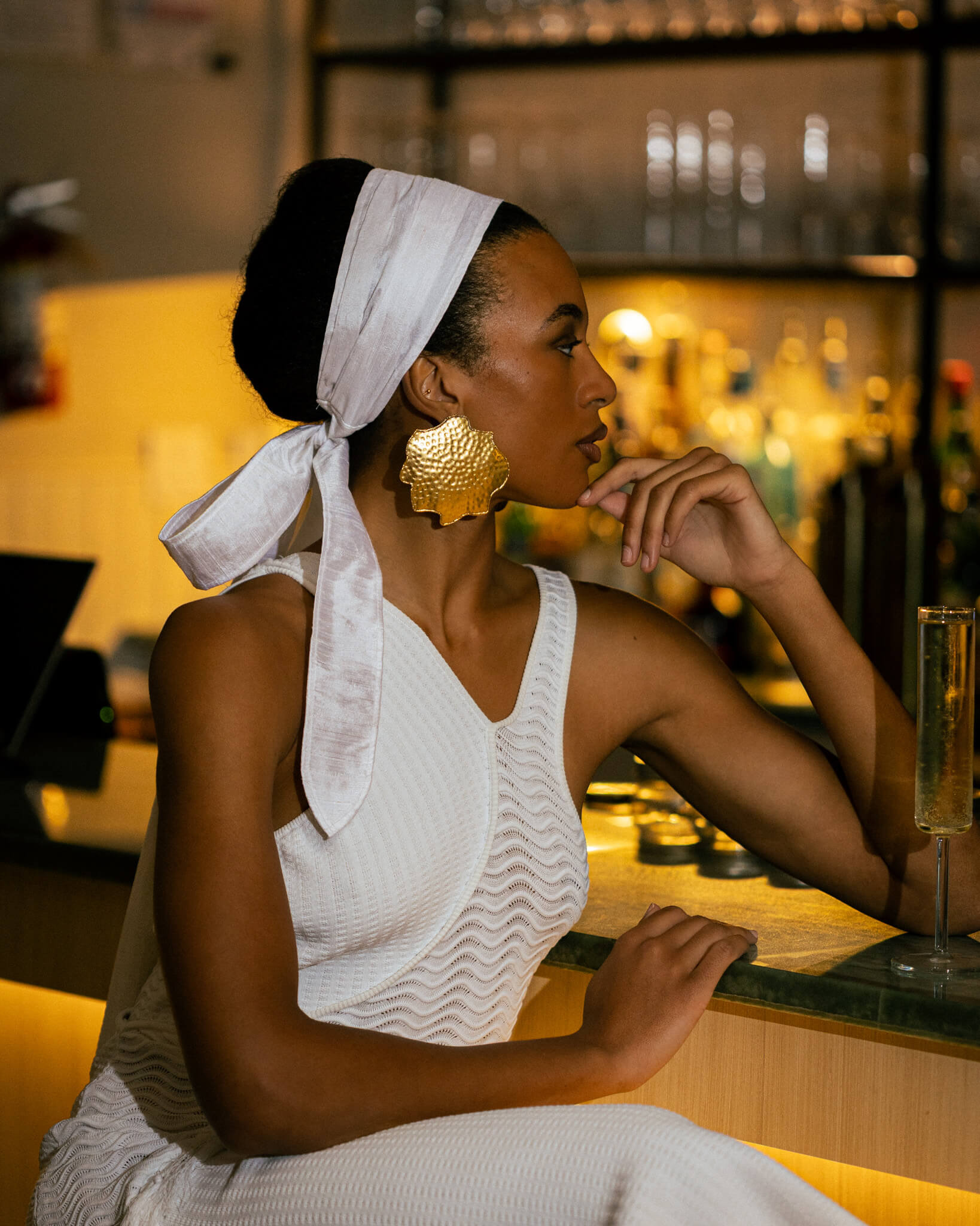 Woman sitting at a bar with a white headscarf and gold earrings.