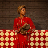 Woman in a red dress with gold accents standing against a brick wall with a checkered floor.
