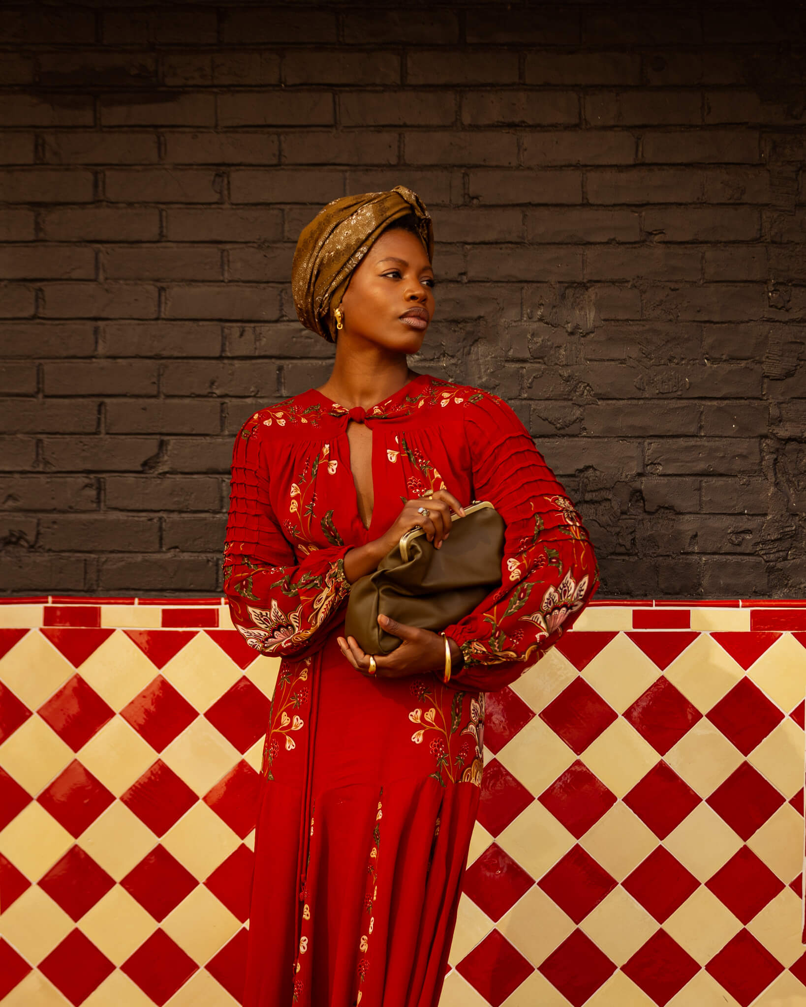 Woman in a red dress with gold accents standing against a brick wall with a checkered floor.
