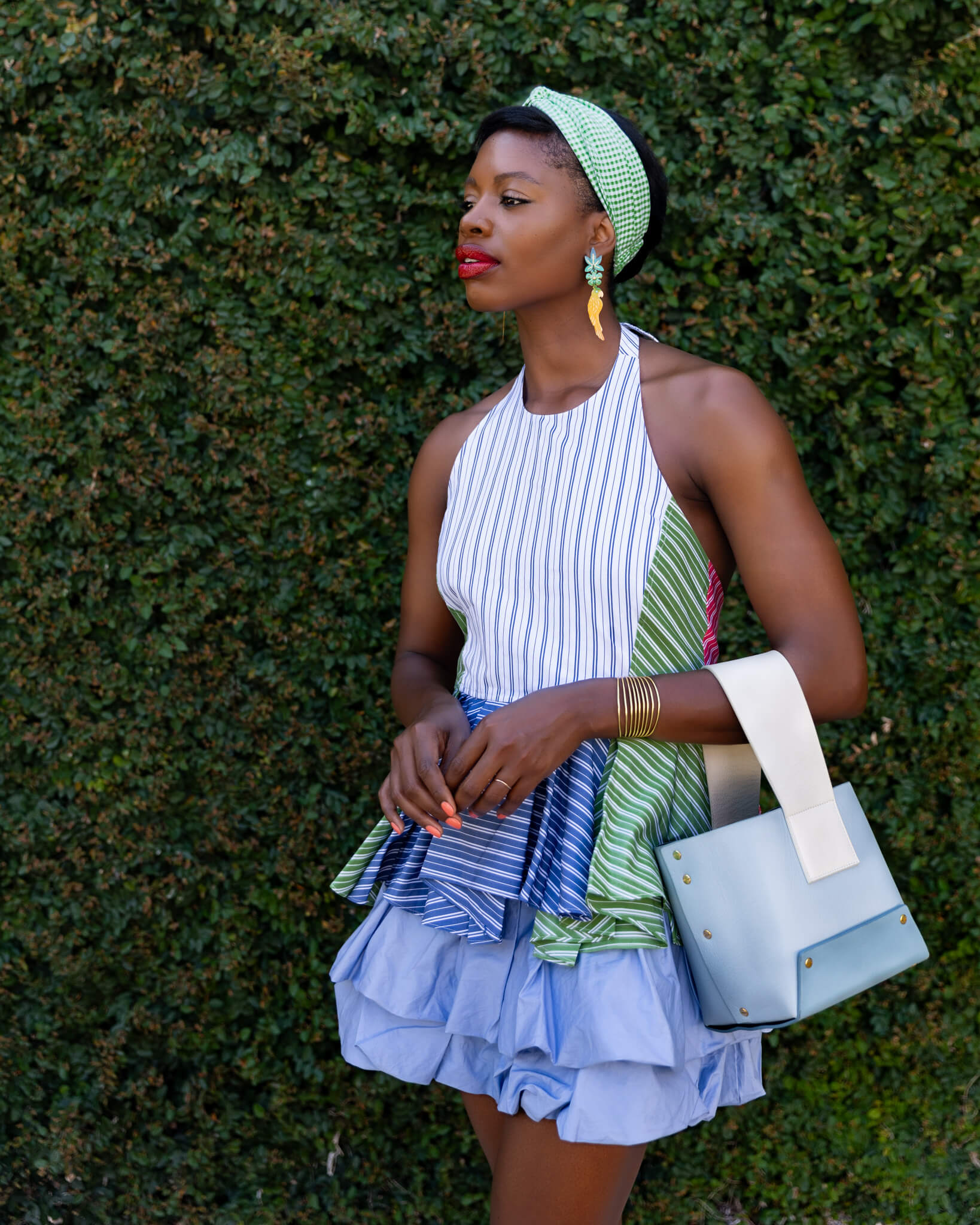 Woman in a stylish outfit with a green headband and blue handbag against a green hedge.