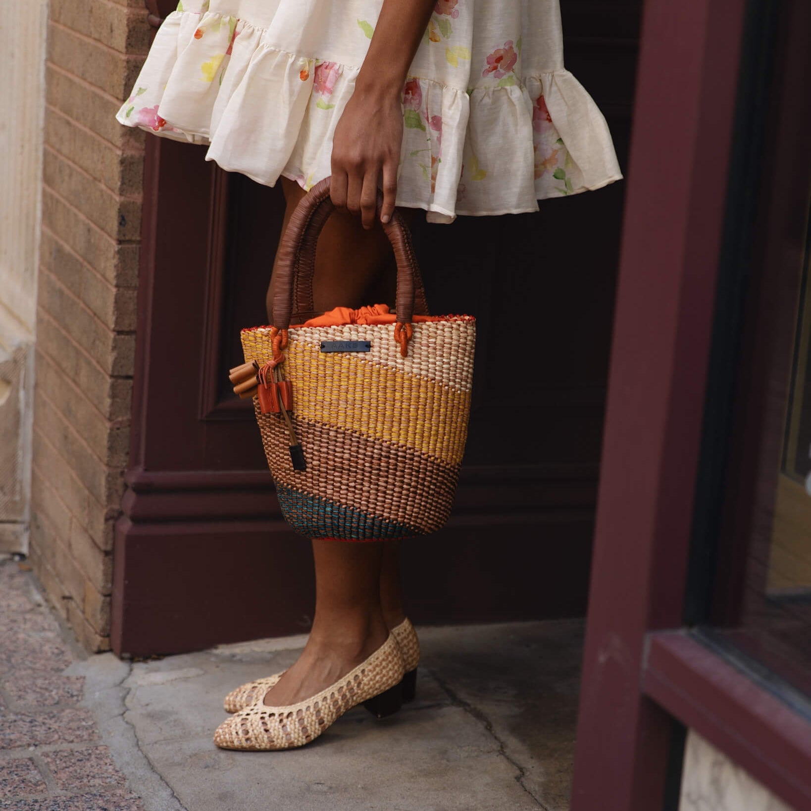 Person wearing a floral dress and holding a colorful woven bag, standing on a sidewalk.