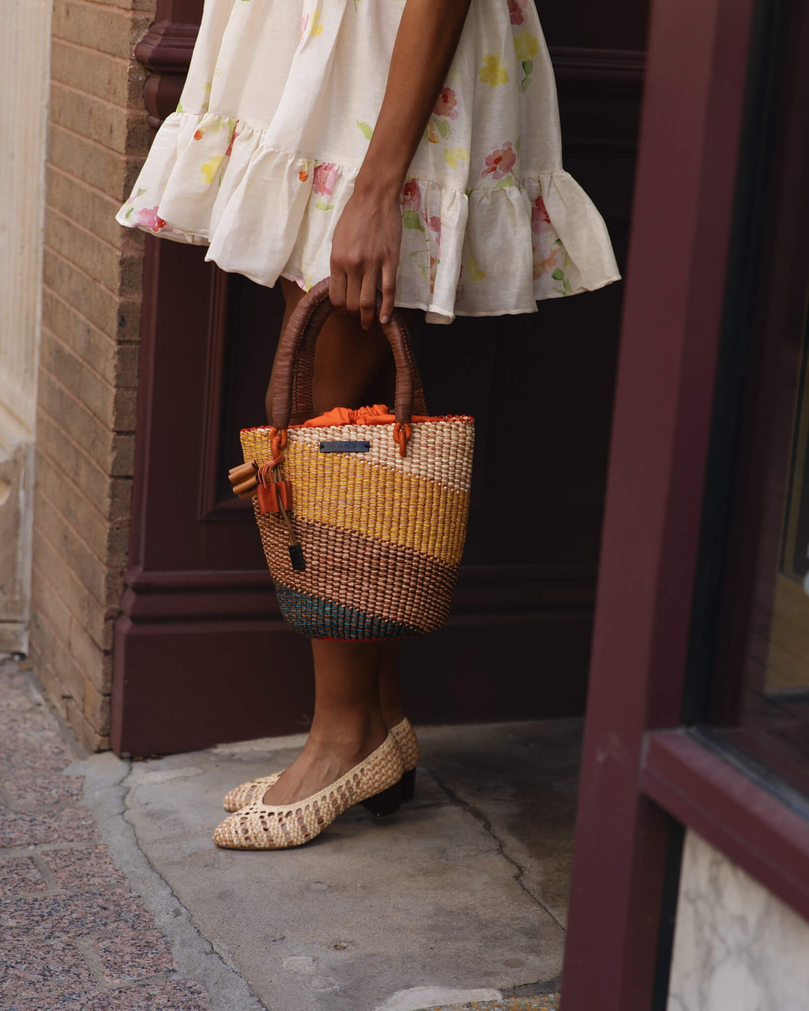 Person wearing a floral dress and holding a colorful woven bag, standing on a sidewalk.