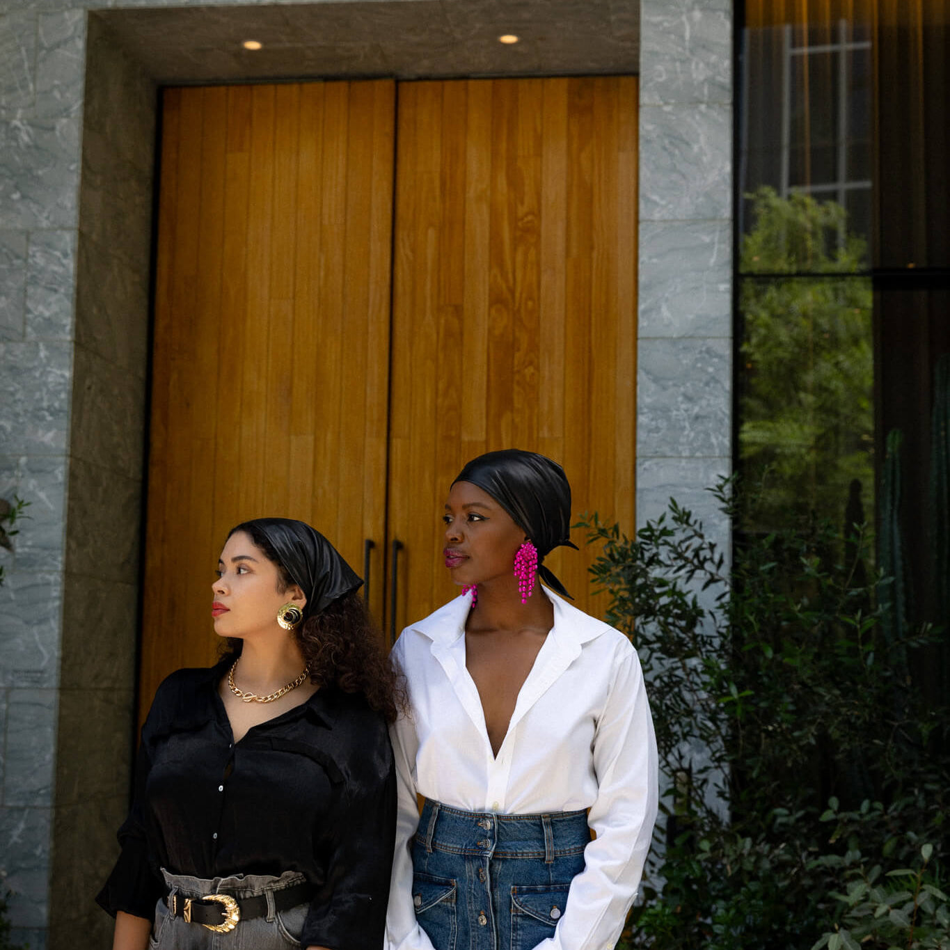 Two women standing outside a building with wooden doors, wearing black  faux leather bandanas